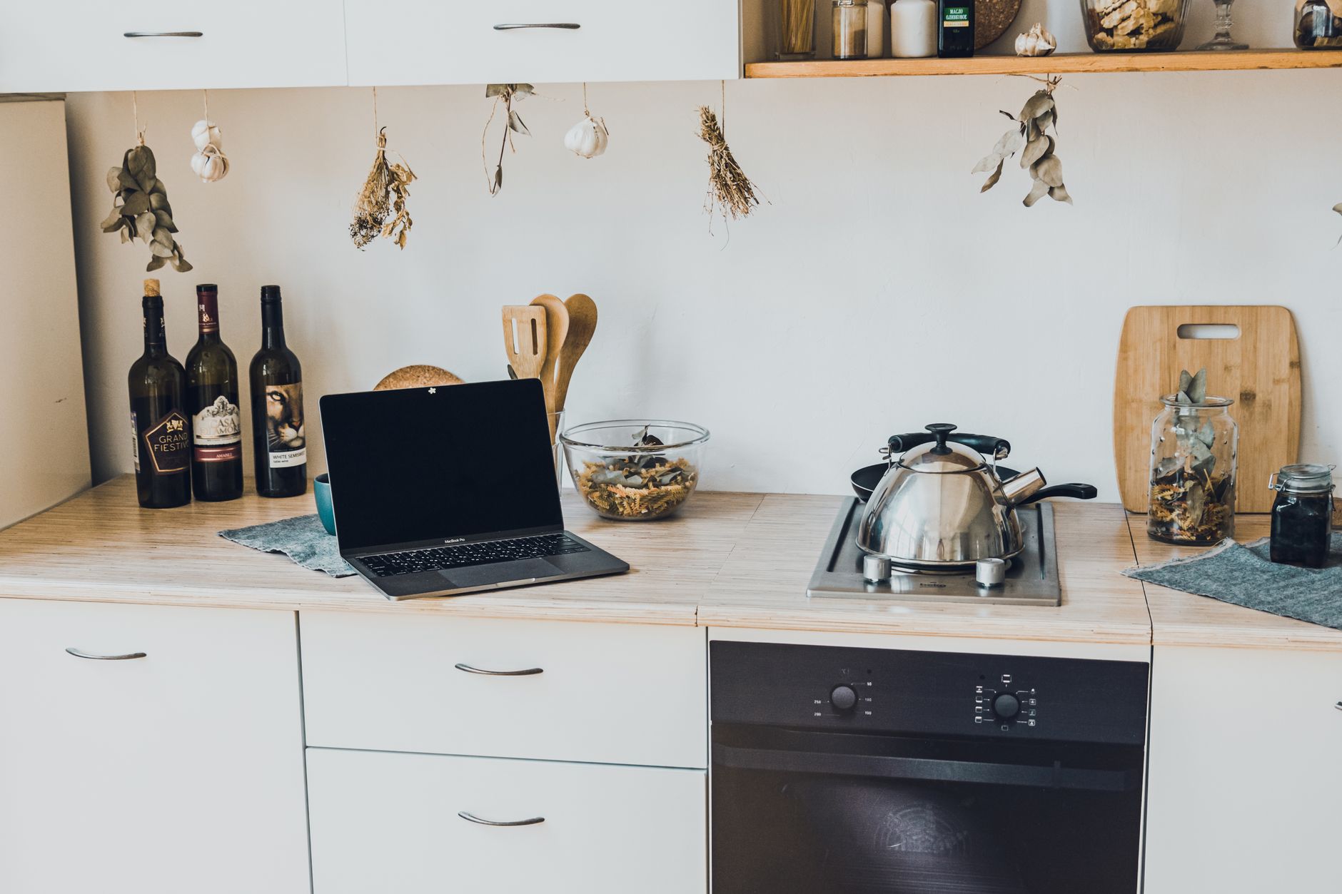 A stylish kitchen interior featuring a laptop, cookware, and decorative elements like hanging herbs.