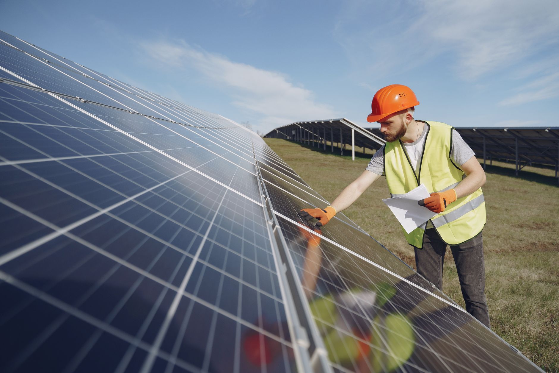 Young bearded male engineer in protective helmet and heat resistant gloves touching solar panel while standing on field with papers on sunny