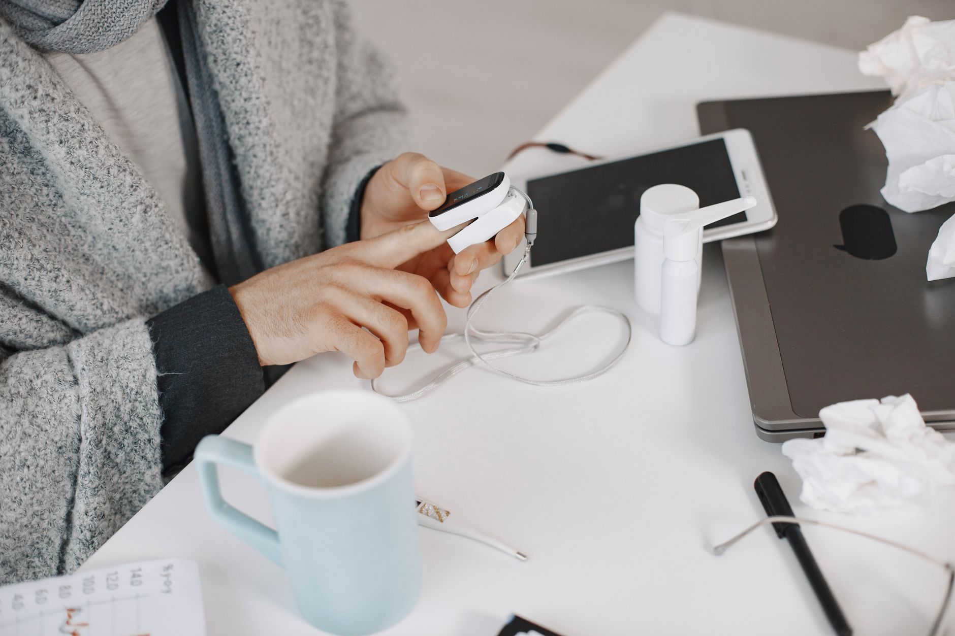 Adult using a pulse oximeter at a home desk, surrounded by tech and healthcare items.