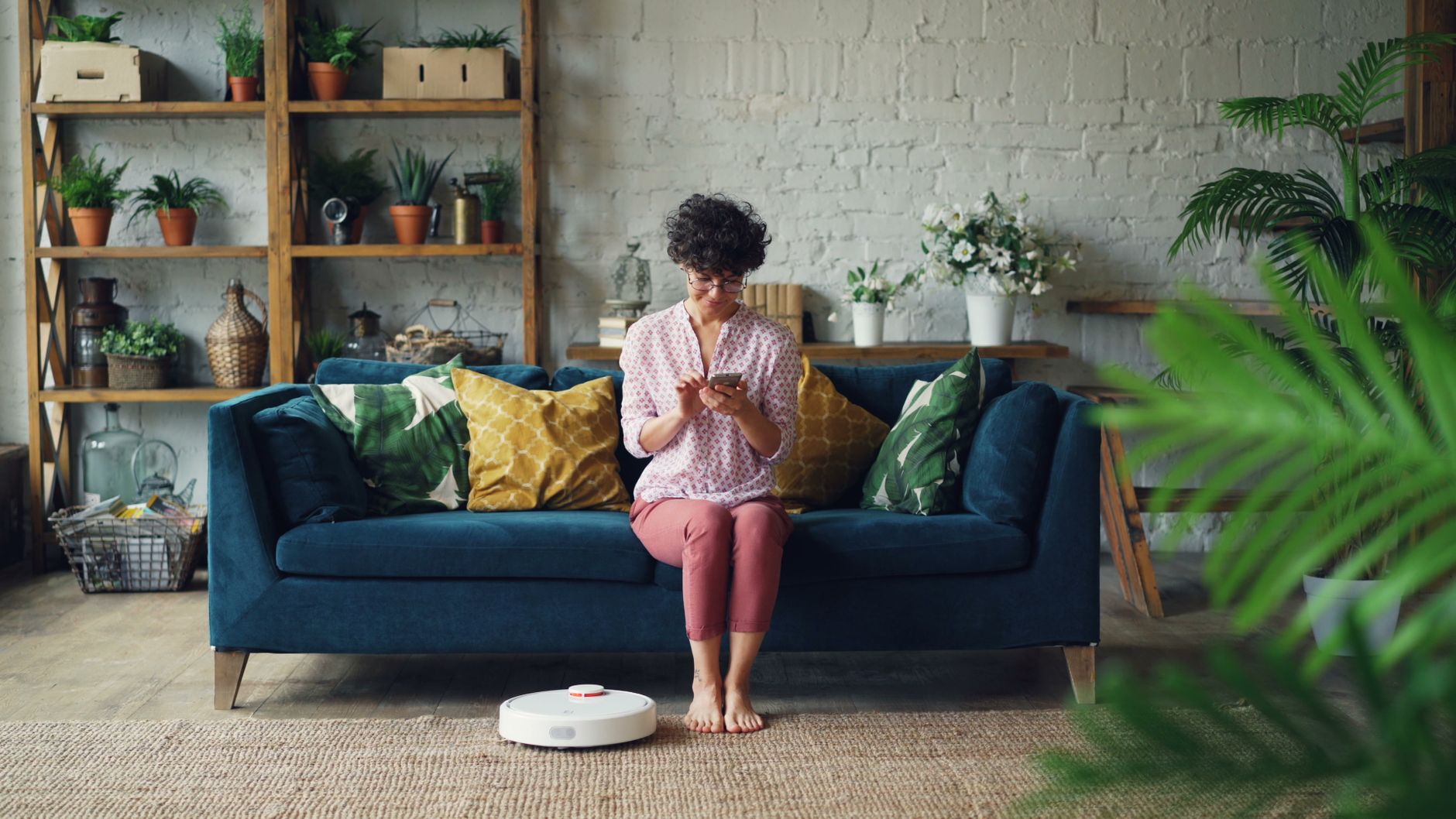 Woman sits on sofa using smartphone while robotic vacuum cleans living room floor.