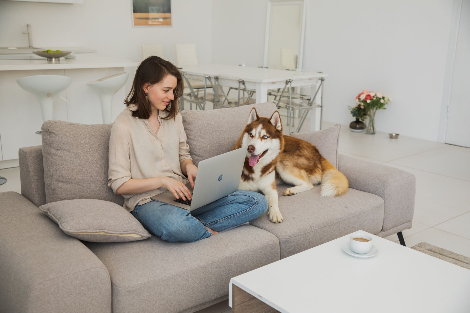 Woman working on laptop with a husky dog beside her on a cozy living room sofa.