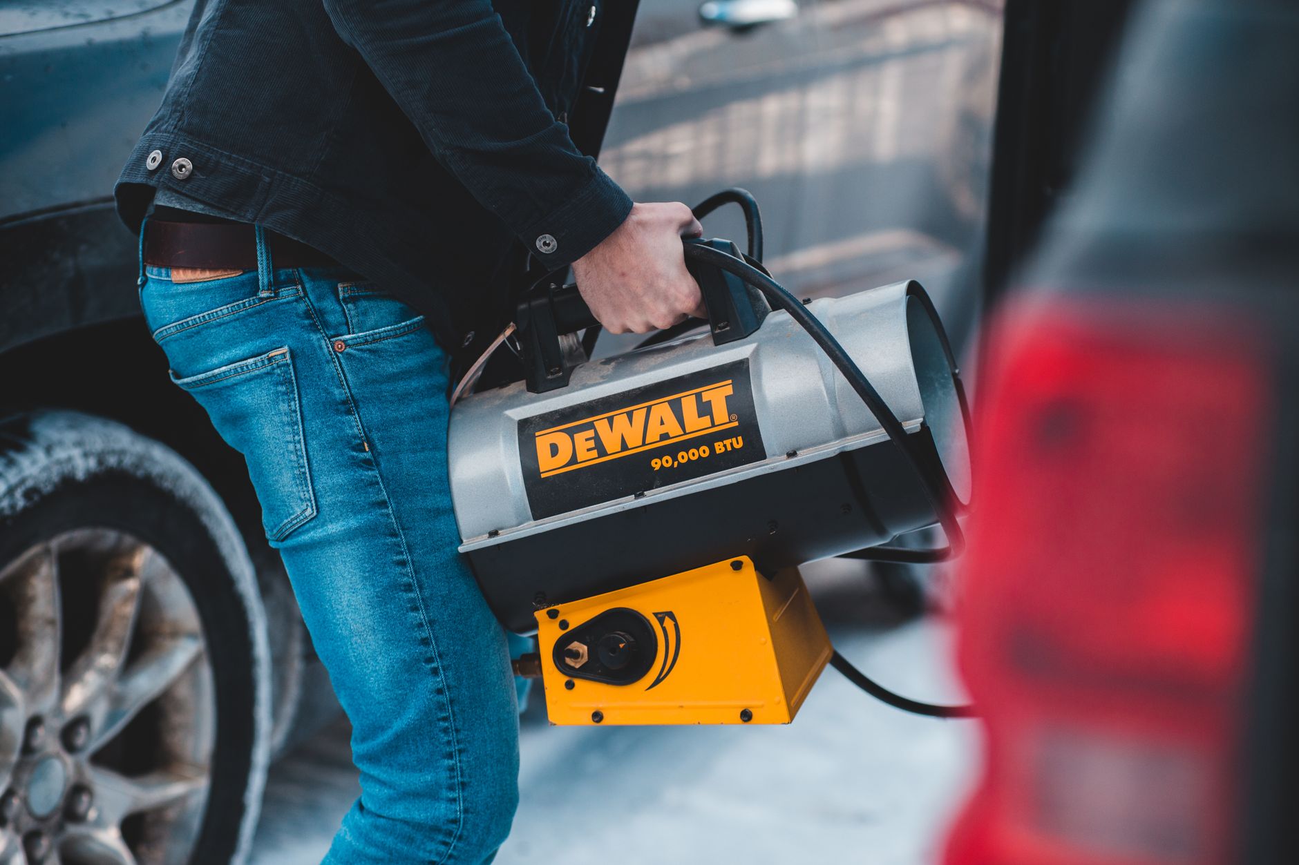 From above side view of crop unrecognizable slim male in jeans carrying portable blow heater with plastic handle and shiny metal tank standi