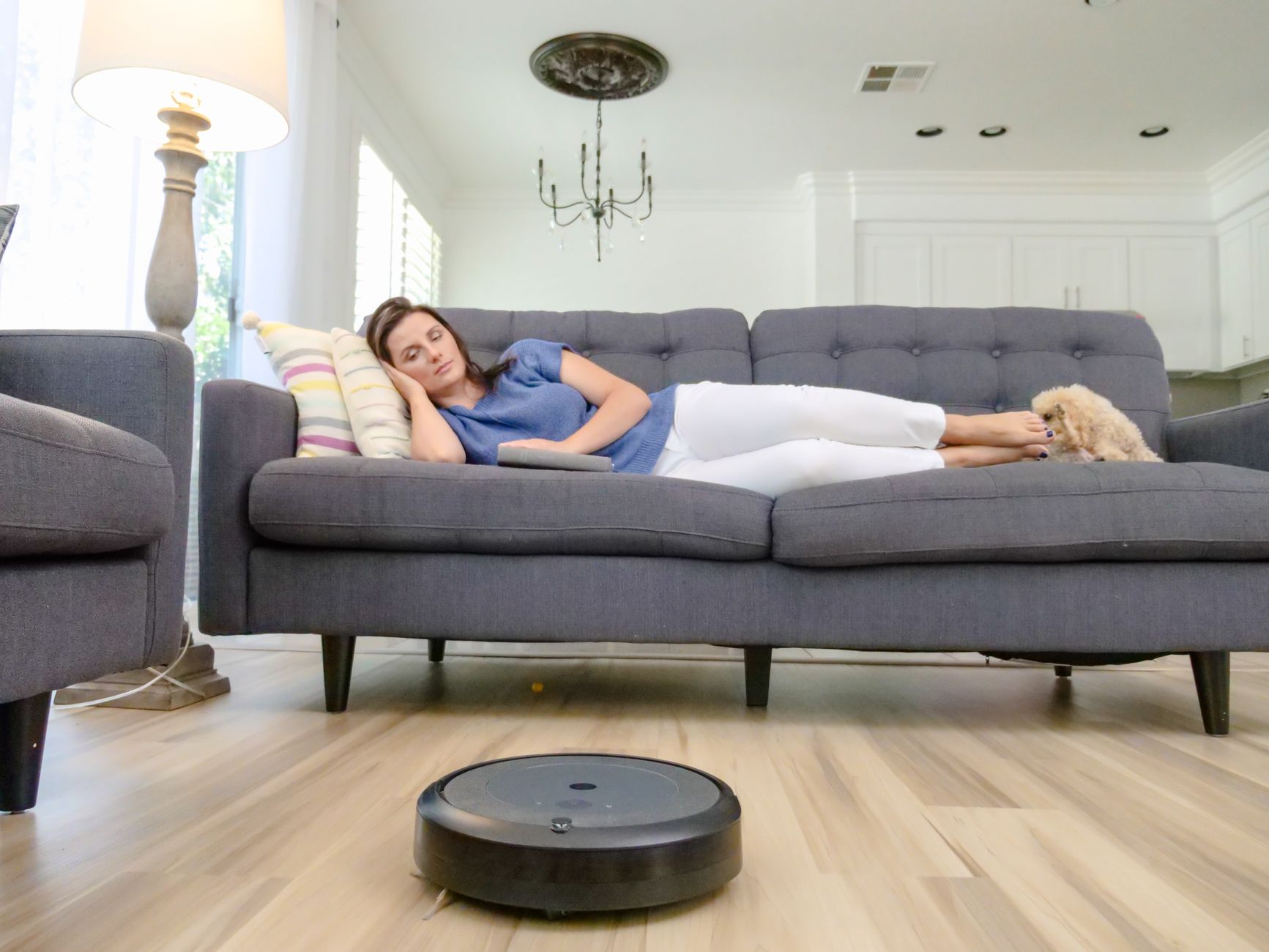 A woman relaxing on a sofa with a robotic vacuum cleaner cleaning the hardwood floor.