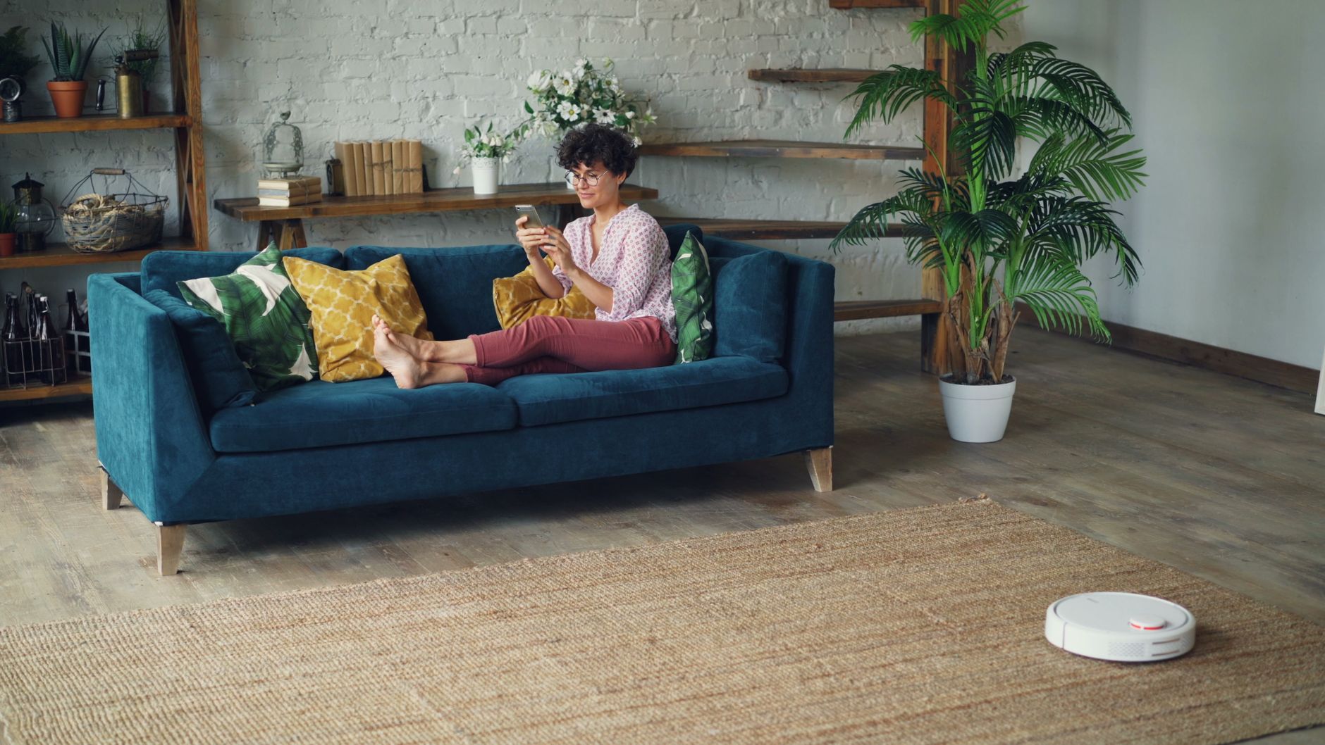 Woman relaxing on a stylish blue sofa at home, using a smartphone with a robotic vacuum on the floor.