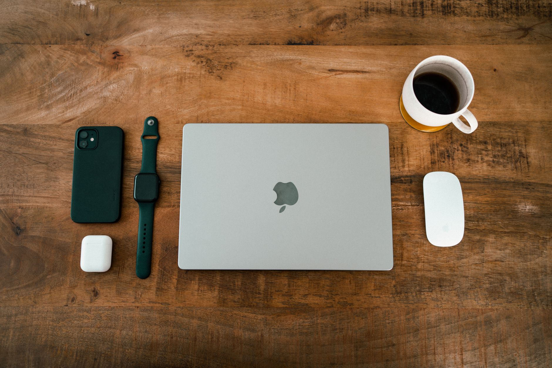 Flat lay image featuring a laptop, smartphone, smartwatch, wireless earbuds, and coffee cup on a wooden desk.