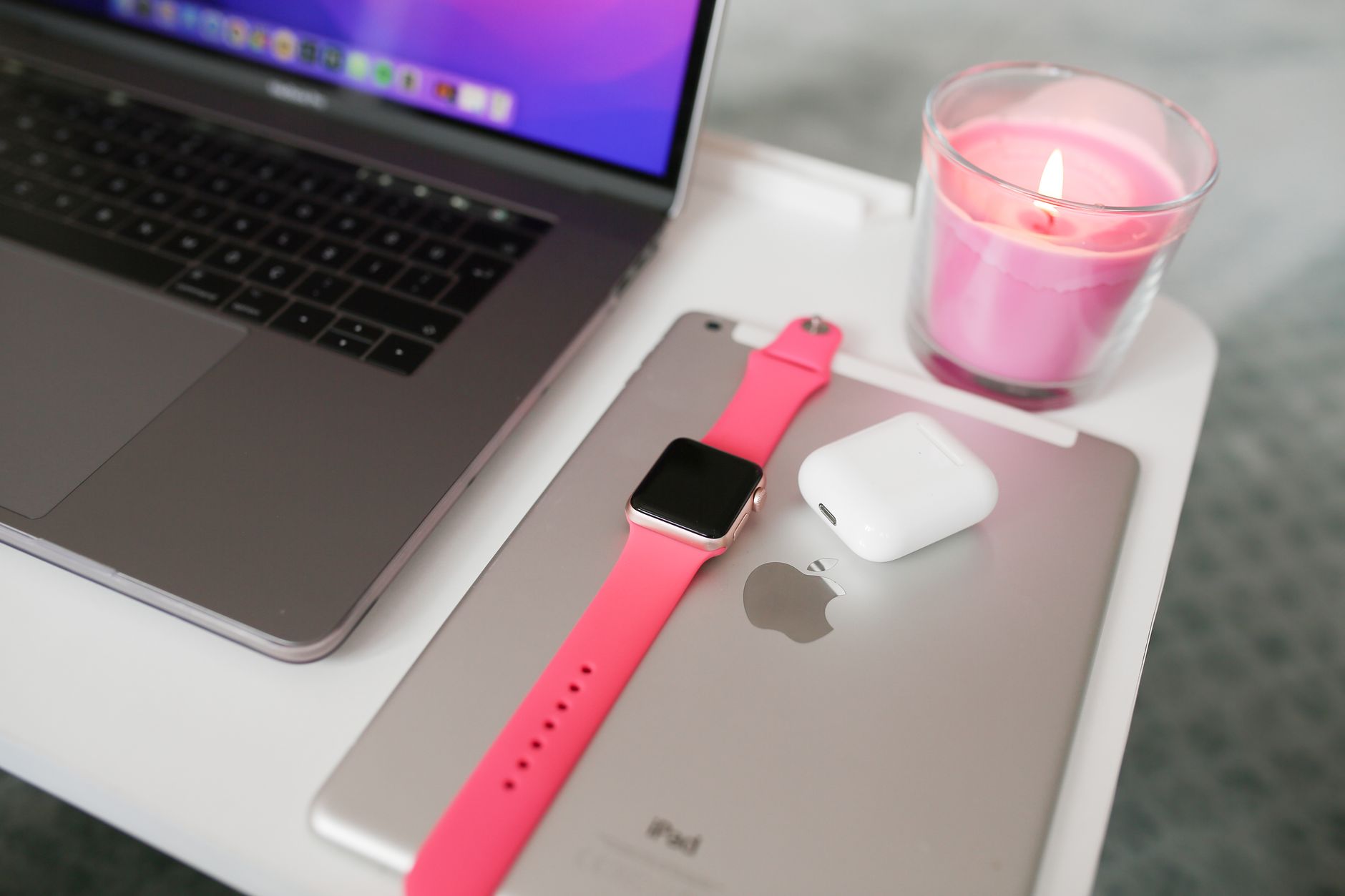 A neatly arranged desk with a laptop, iPad, smartwatch, AirPods, and candle.