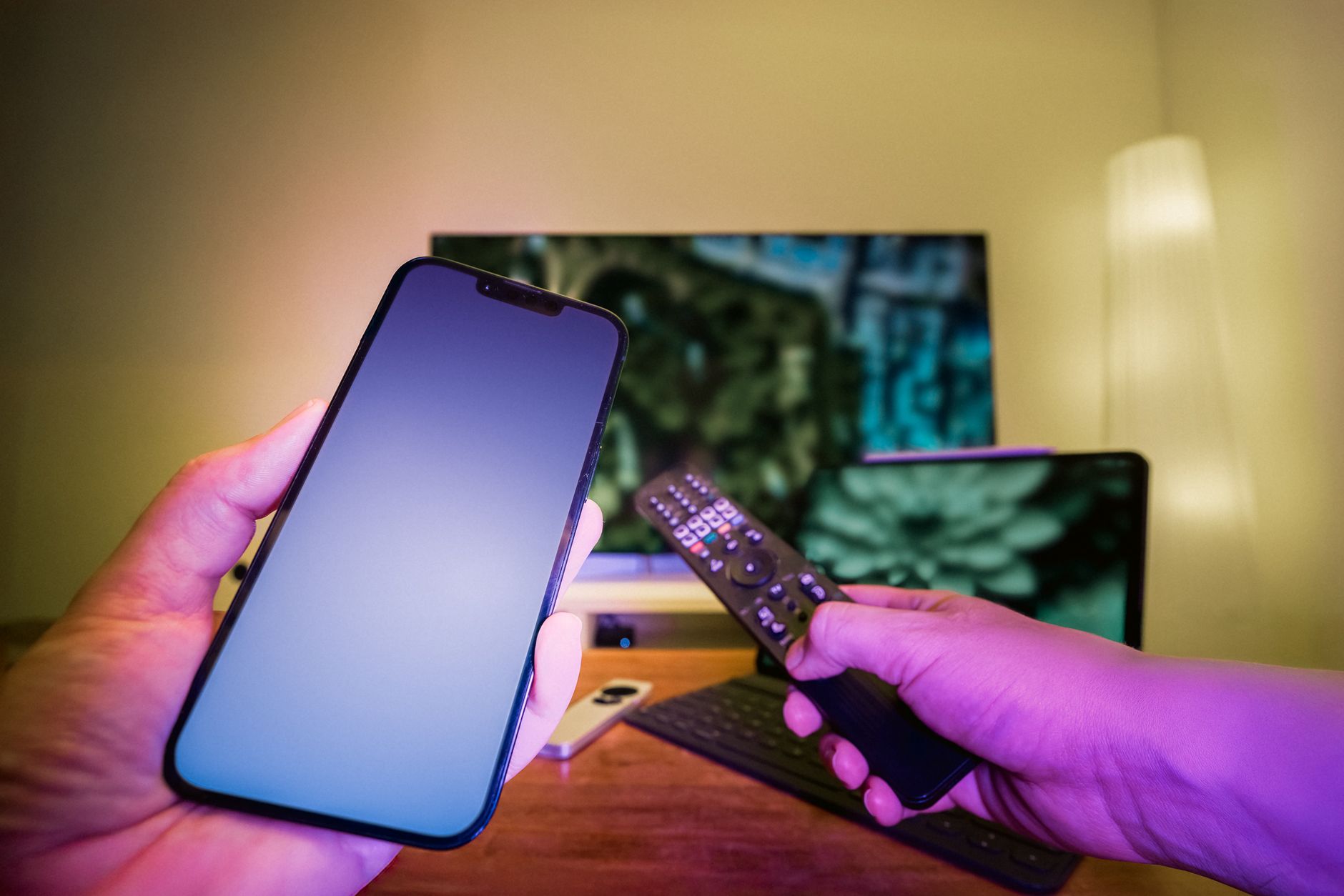 Hands holding a smartphone and remote control in a high-tech home setup with TV and other electronic devices.