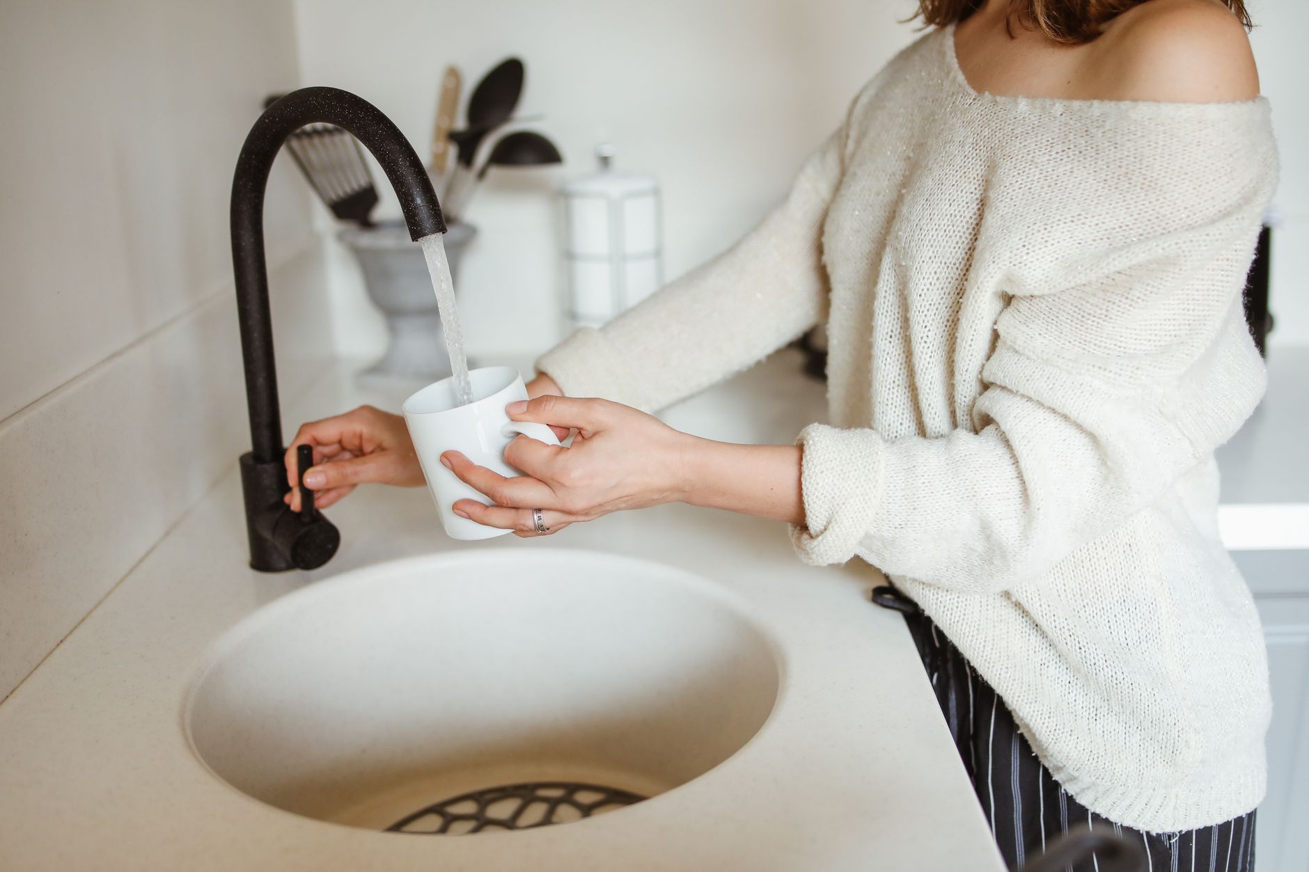 A woman filling a white cup with water from a black faucet in a modern kitchen.